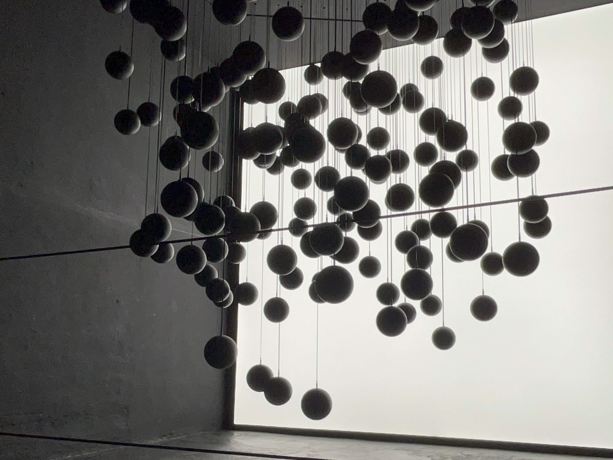 Dozens of black spheres hanging from the ceiling at various heights and depths. Walls, ceiling, and floor are made of concrete. A white screen contrasts with the spheres in the background.