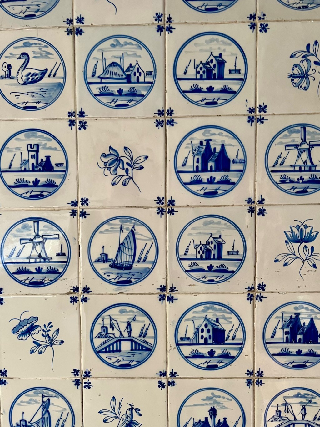 White ceramic tiles with blue motifs. Various landscapes are depicted (mill, boat, house), each within a blue circle.