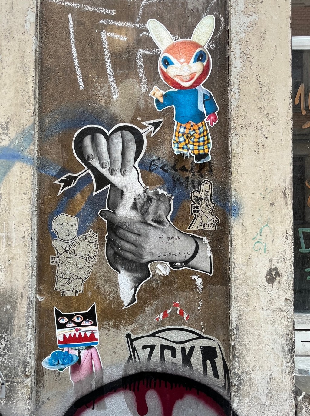 Graffiti and stickers on a street wall. A hand feeds a mall animal. Cartoonish and colourful figures of anthropomorphic animals . Wall is brownish-grey